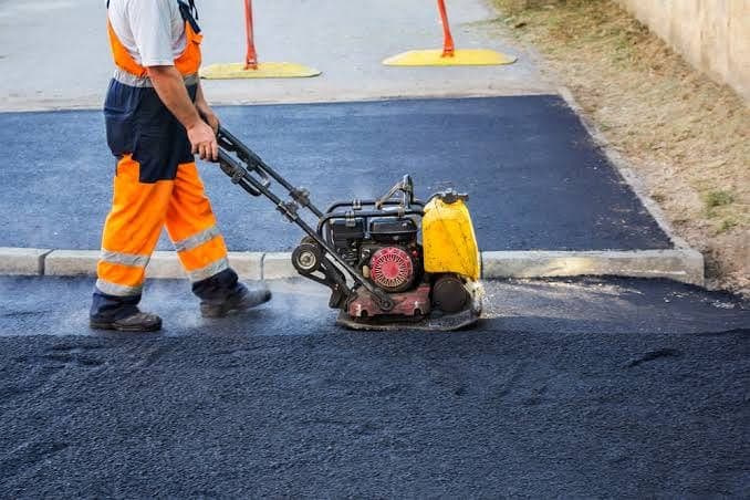 Worker in orange hi-vis work pants running a plate compactor over fresh asphalt next to a concrete curb