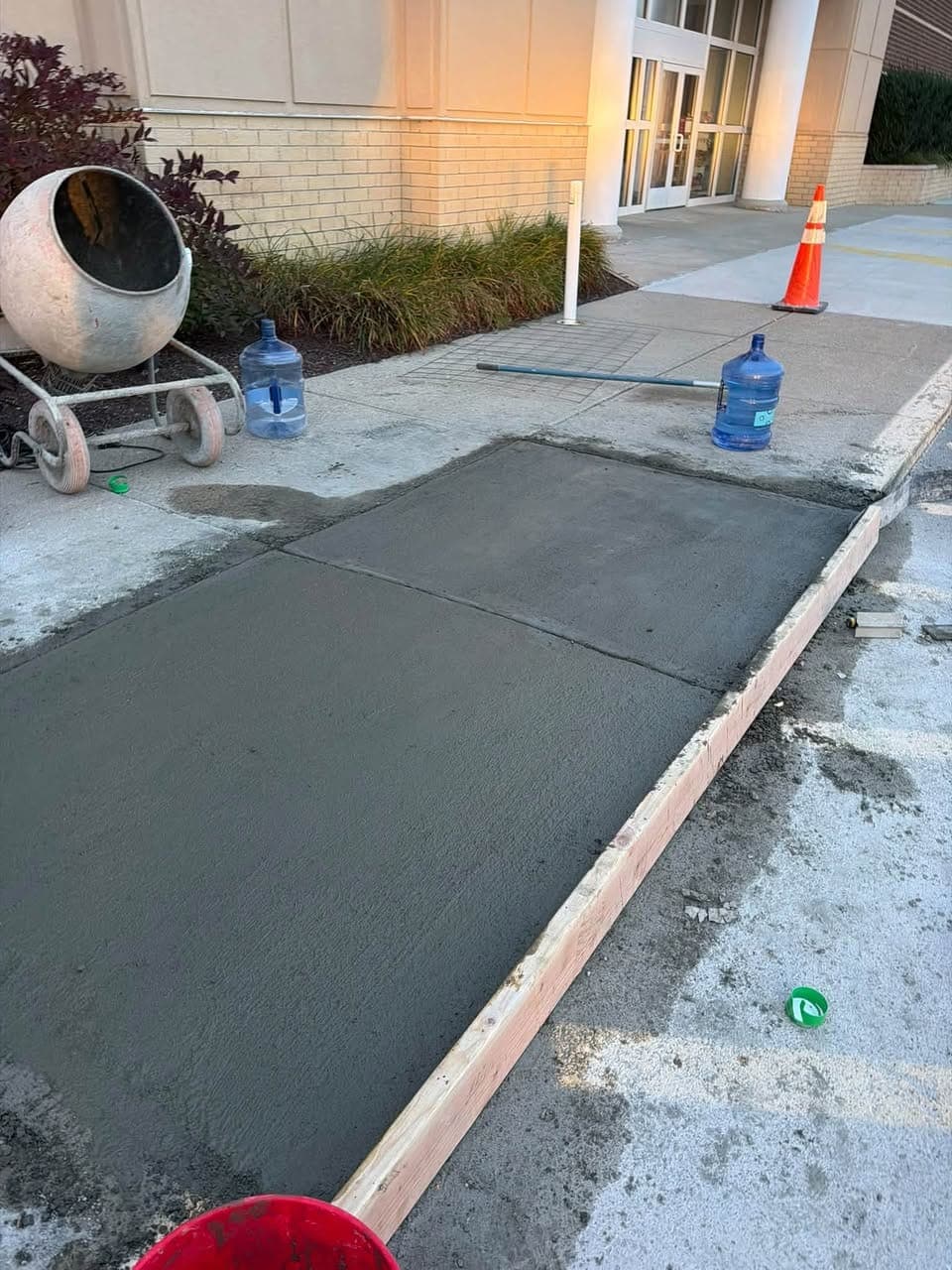 Freshly poured concrete sidewalk panels with a wood form along the curb outside a commercial building, cement mixer at left