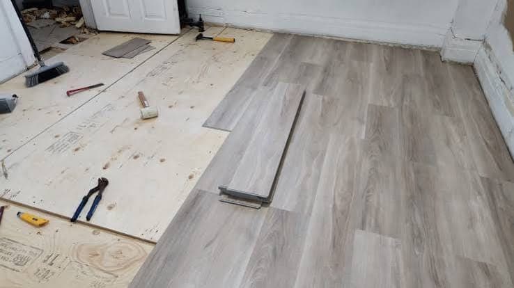 Gray wood-look vinyl plank flooring being laid over a plywood subfloor, with a pull bar, mallet and hand tools