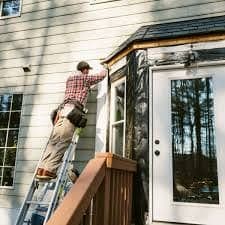 Worker in a plaid shirt and tool belt on an extension ladder, working on exterior trim beside a glass door