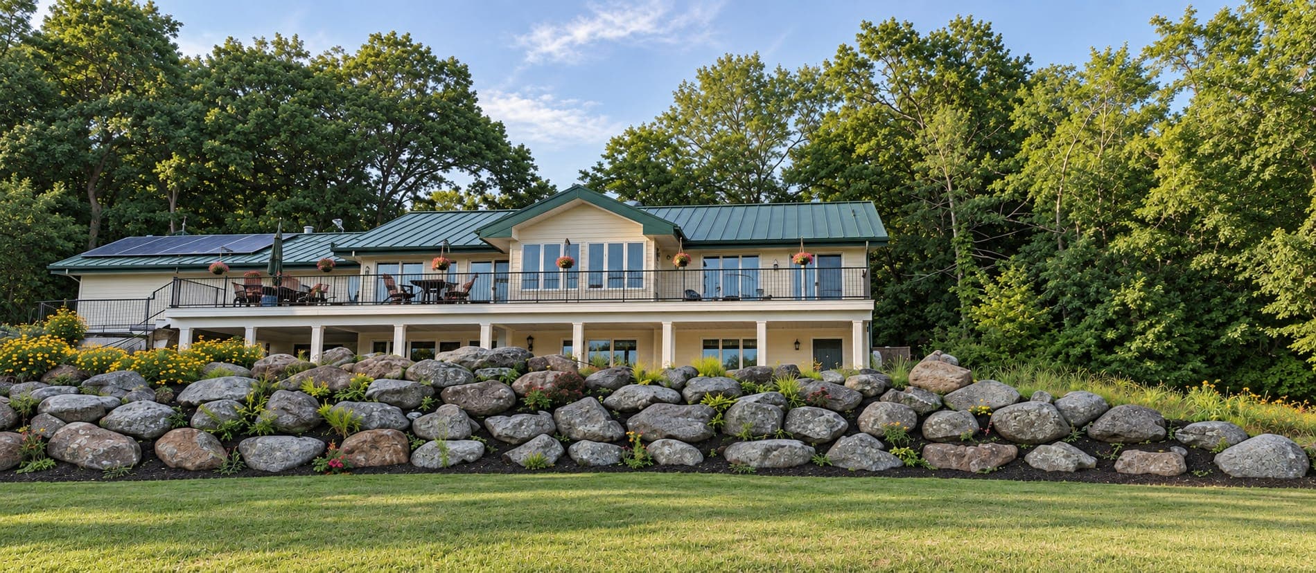 Planted boulder retaining wall and lawn below a cream house with a green metal roof, solar panels and deck